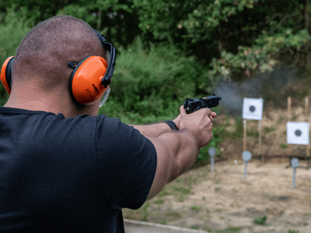 Atirador usando proteção auricular e ocular enquanto aponta uma pistola para alvos posicionados em um estande de tiro.
